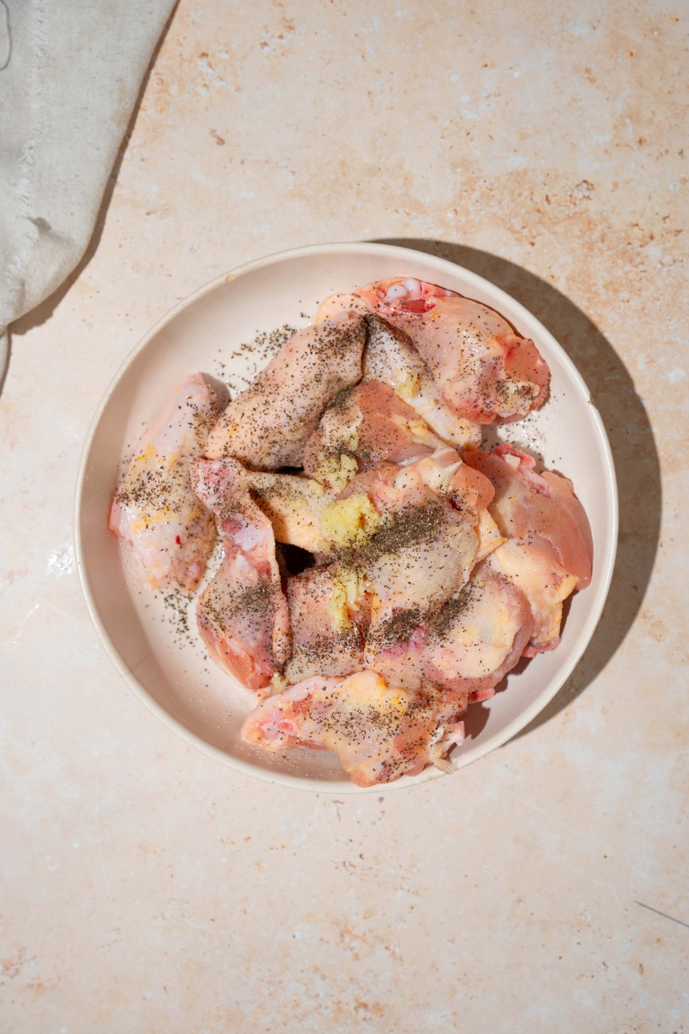 A white plate with raw chicken wings topped with seasonings. The plate is on a tan counter.