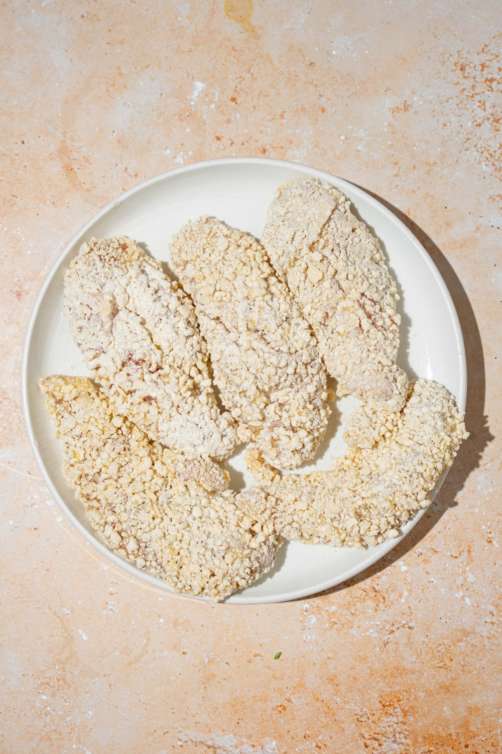 A white plate with breaded uncooked chicken strips. The plate is on a tan counter.