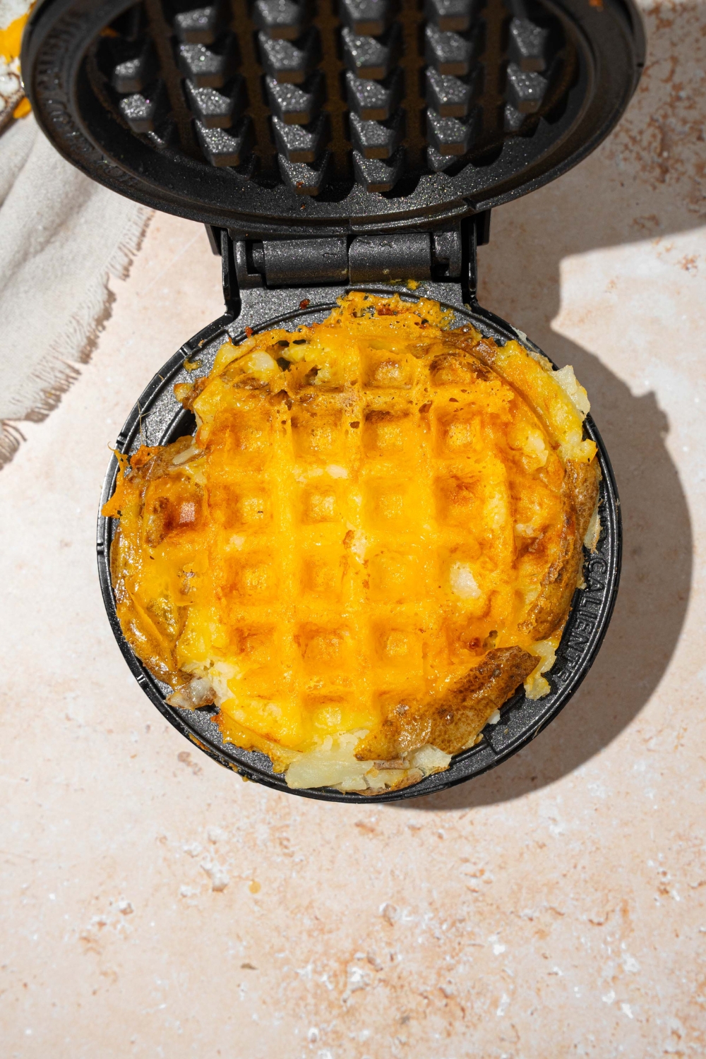 An open waffle maker with a cooked crispy potato waffle on the inside. The waffle maker is on a tan counter.