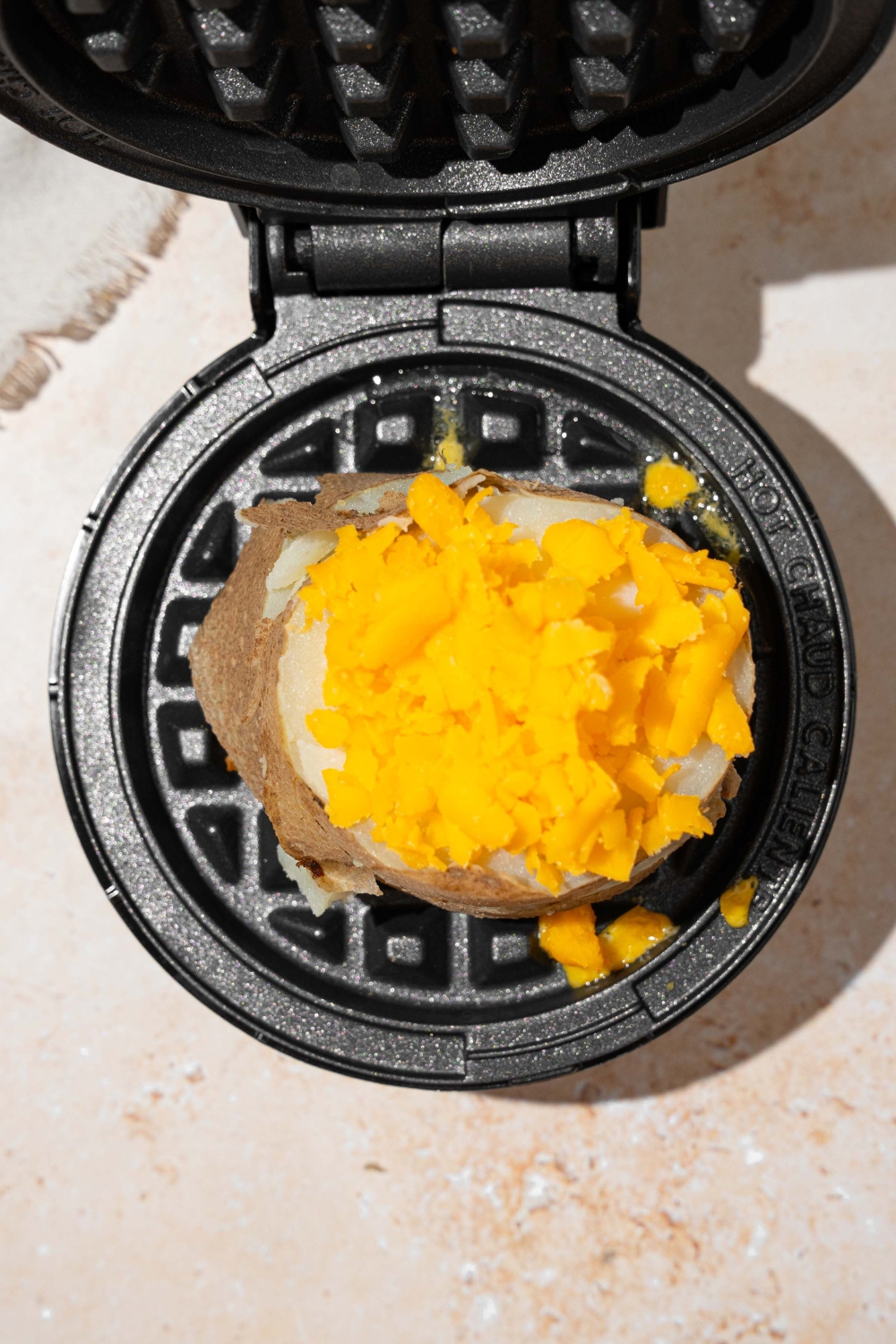 An open waffle maker with fresh potato topped with shredded cheese. The waffle maker is on a tan counter.
