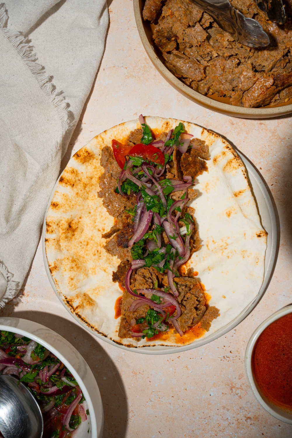 A white plate with an open doner kebab over pita bread topped with roasted vegetables and fresh parsley. The plate is on a tan counter with a plate of kebab meat with a white cloth napkin.