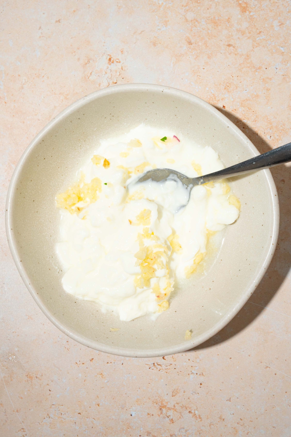 A large white bowl with a spoon mixing a yogurt and garlic sauce. The bowl is on a tan counter.