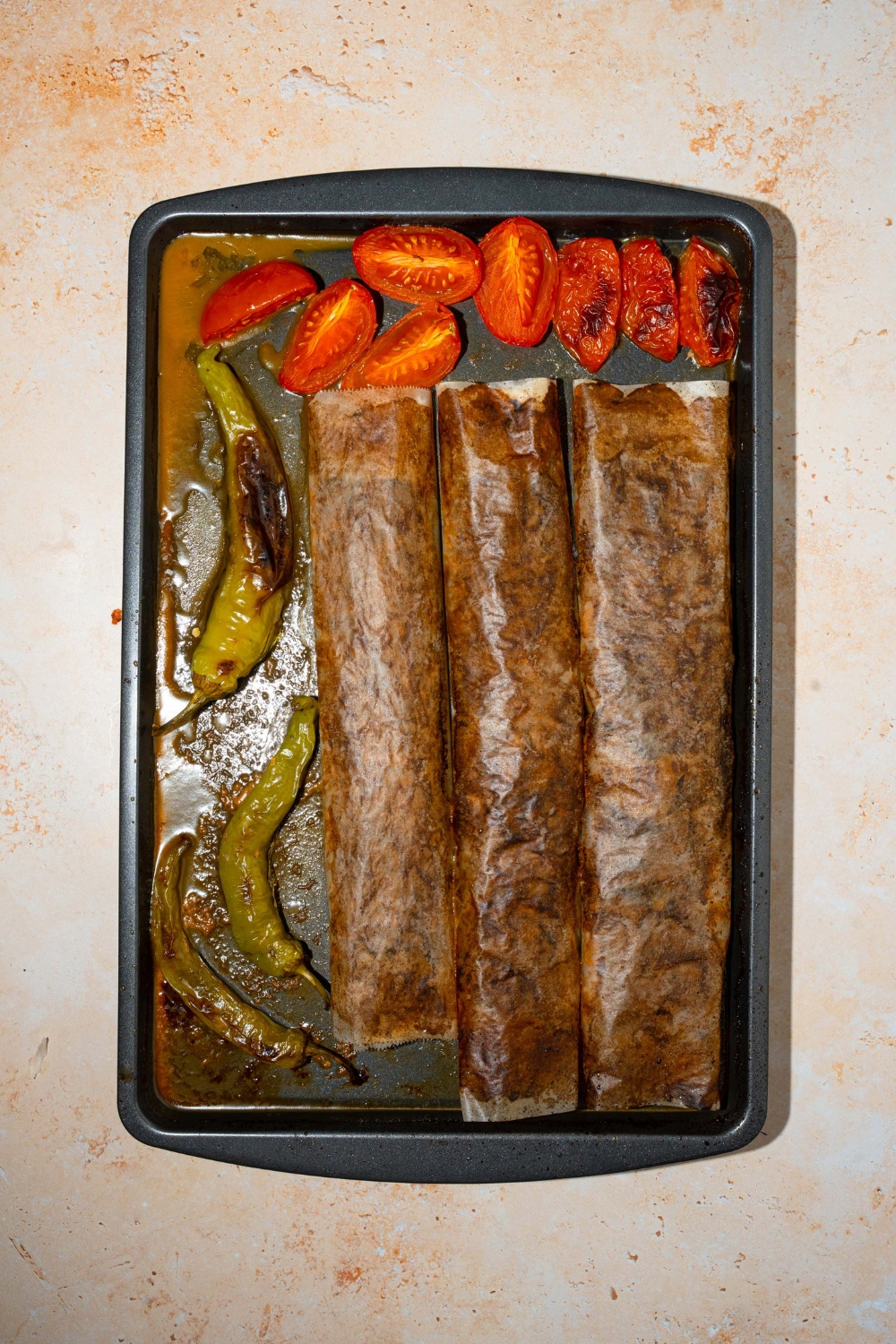 A baking sheet with three wrapped doner kebab meat logs with roasted green peppers and tomatoes. The sheet is on a tan counter.