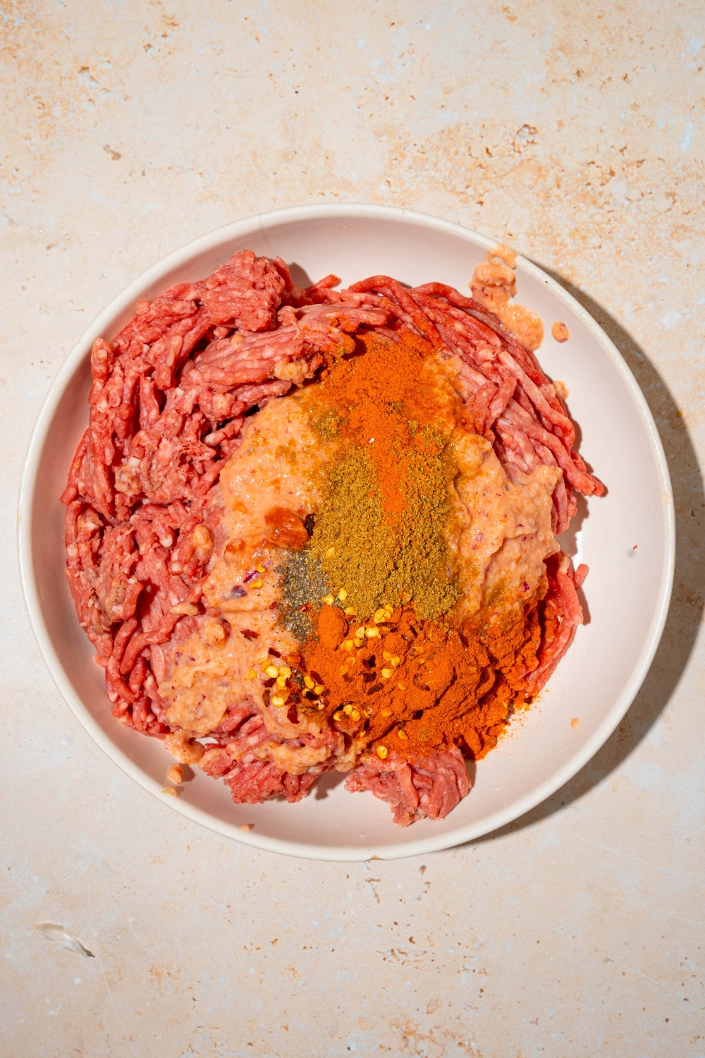 A white bowl with ground beef topped with ground cumin, paprika, red pepper flakes, salt, and pepper. The bowl is on a tan counter.