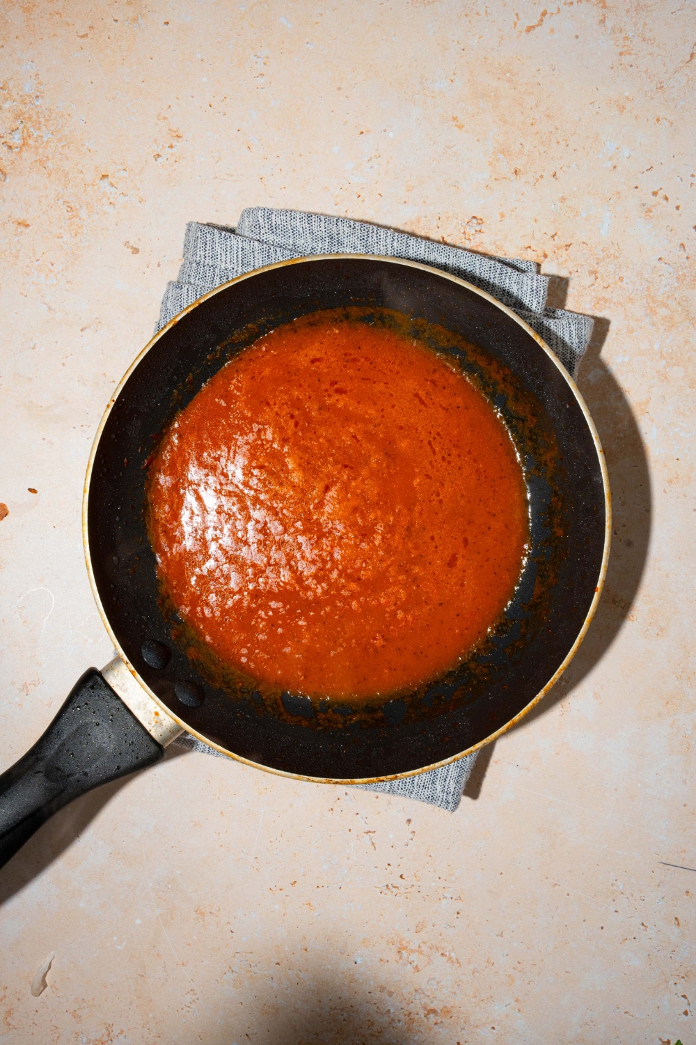 A skillet with tomato paste cooking with butter and water. The skillet is on a tan counter with a white cloth napkin.