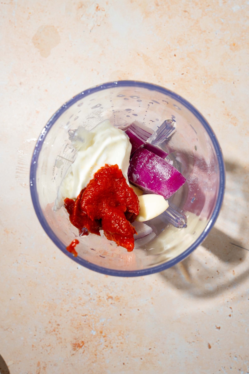 A food processor jar with yogurt, onion, garlic, and tomato paste. The jar is on a tan counter.