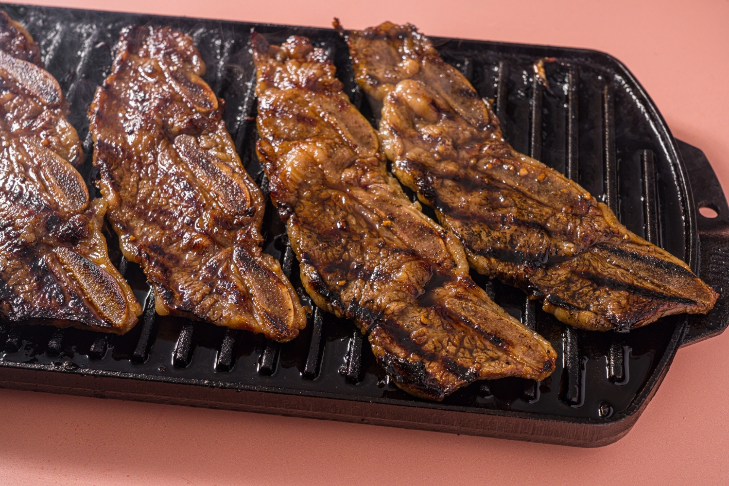 A cast iron grill with several marinated flanken short ribs. The grill is on a pink counter.