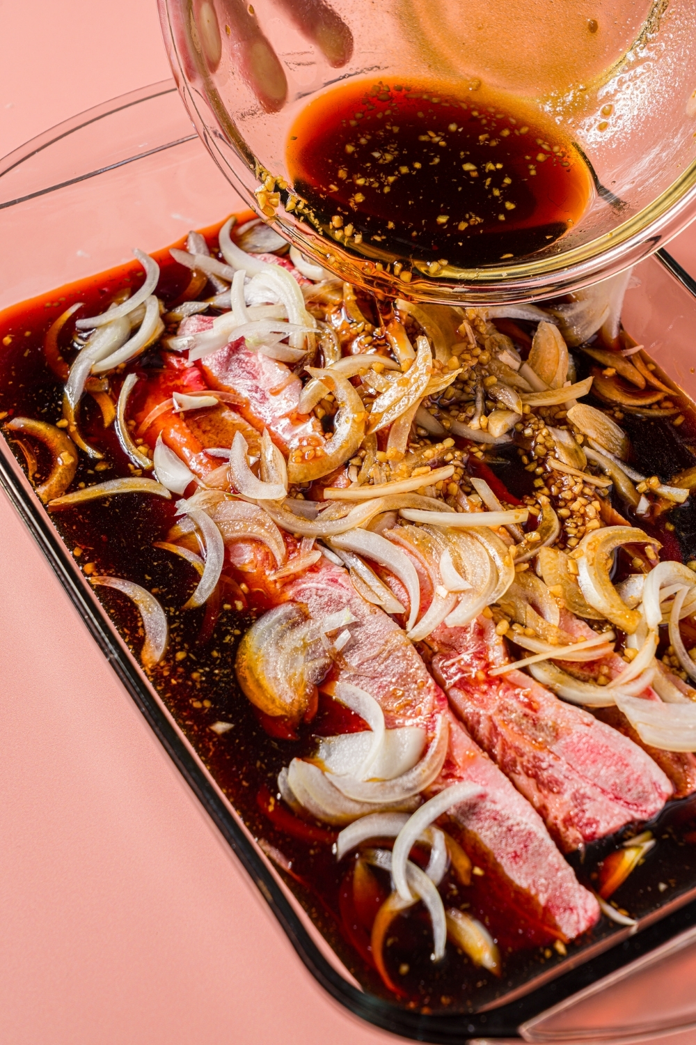 A glass baking dish with uncooked flanken short ribs and sliced onions. A bowl of marinade is being poured over the ribs.
