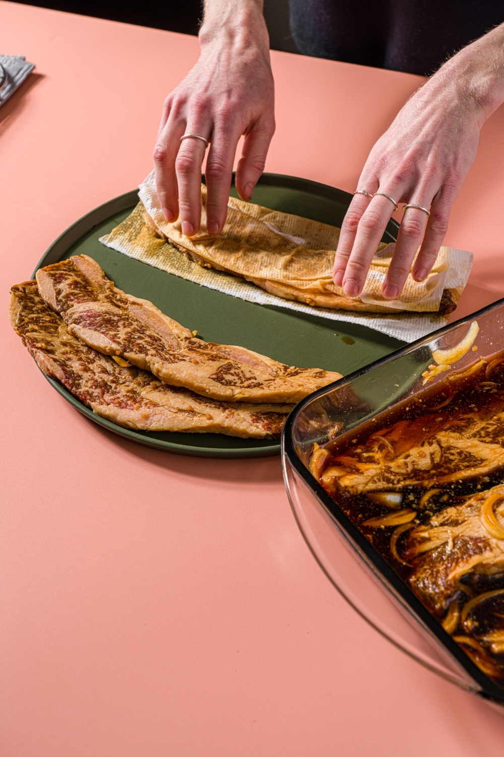A green plate with flanken beef short ribs being wrapped in paper towel and patted dry. The plate is on a pink counter.