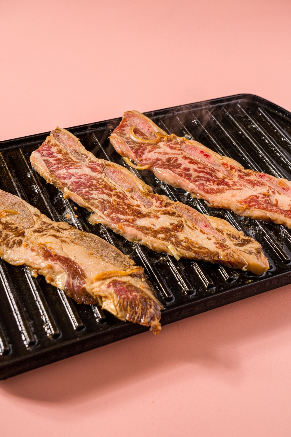 A cast iron grill with several marinated flanken beef short ribs cooking. The grill is on a pink counter.