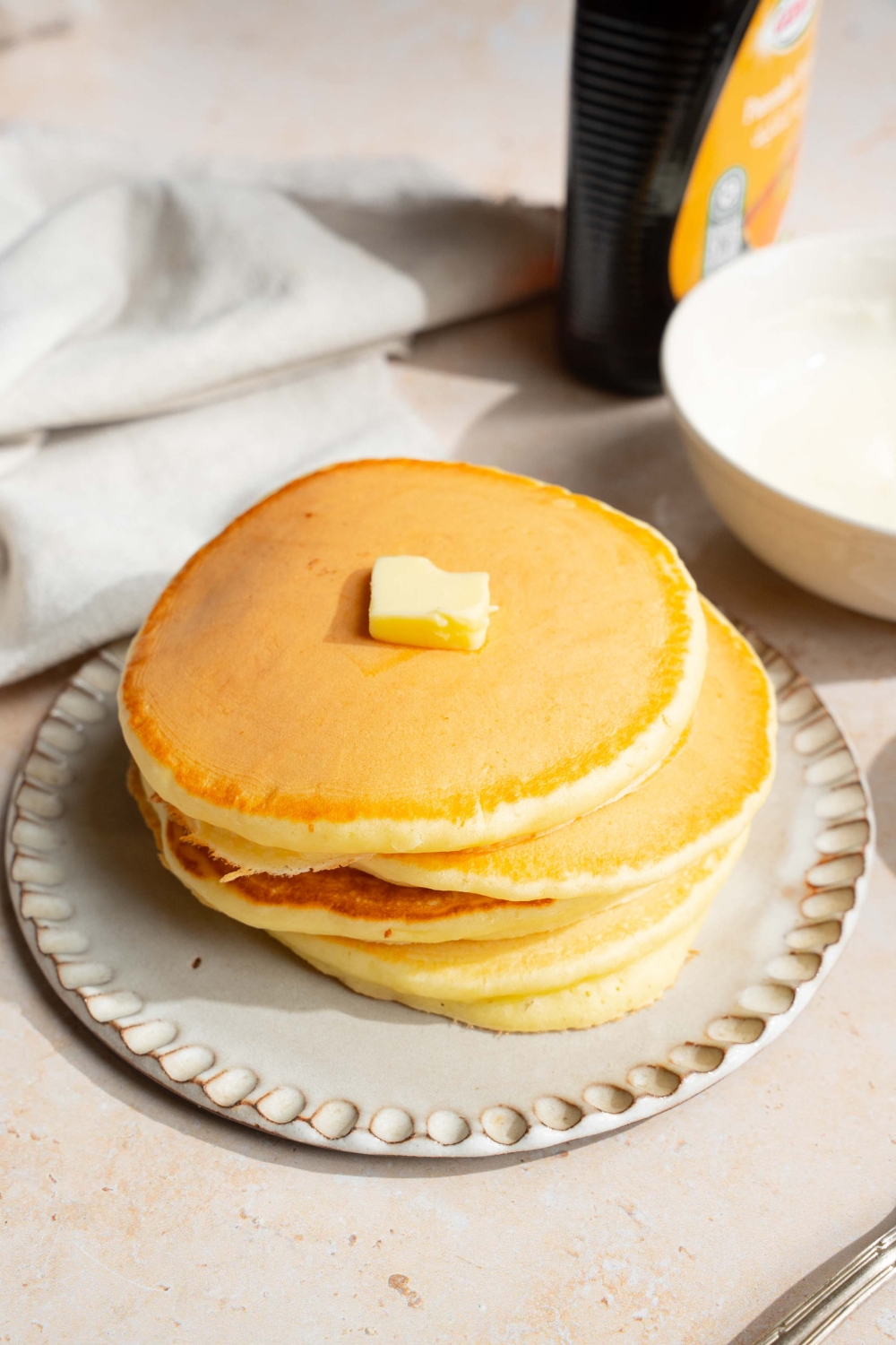 A white plate with a stack of pancakes with self rising flour topped with a pat of butter. The plate is on a tan counter with a jar of syrup and a white cloth napkin.