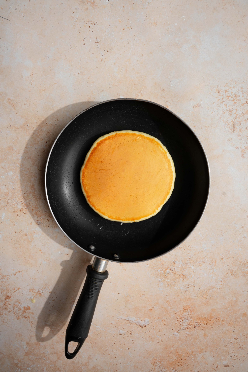 A skillet with a flipped pancake with self rising flour. The skillet is on a tan counter.
