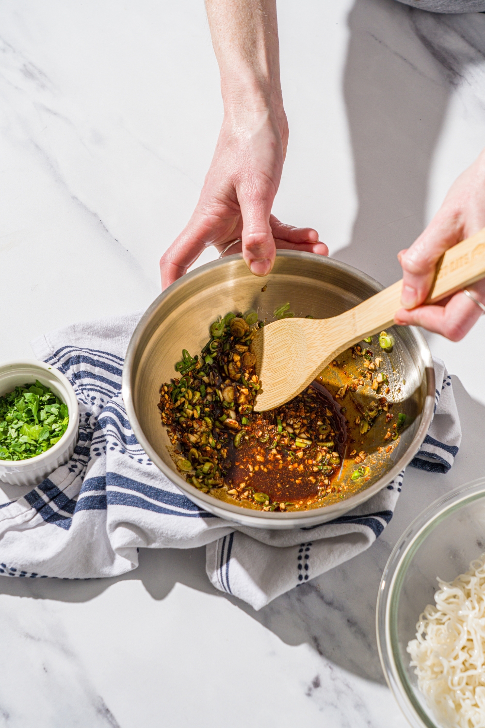 A wooden spoon mixing seasonings and sauces to make a chili sauce. The bowl is on a marble counter with a white cloth napkin and small bowl of sliced green onions.