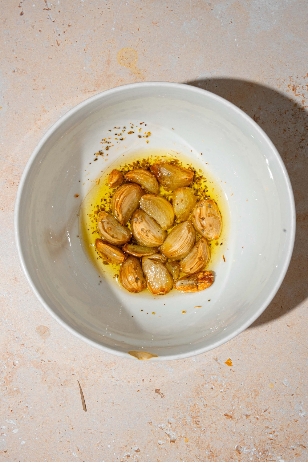 A white bowl with roasted garlic in seasonings and oil. The bowl is on a tan counter.
