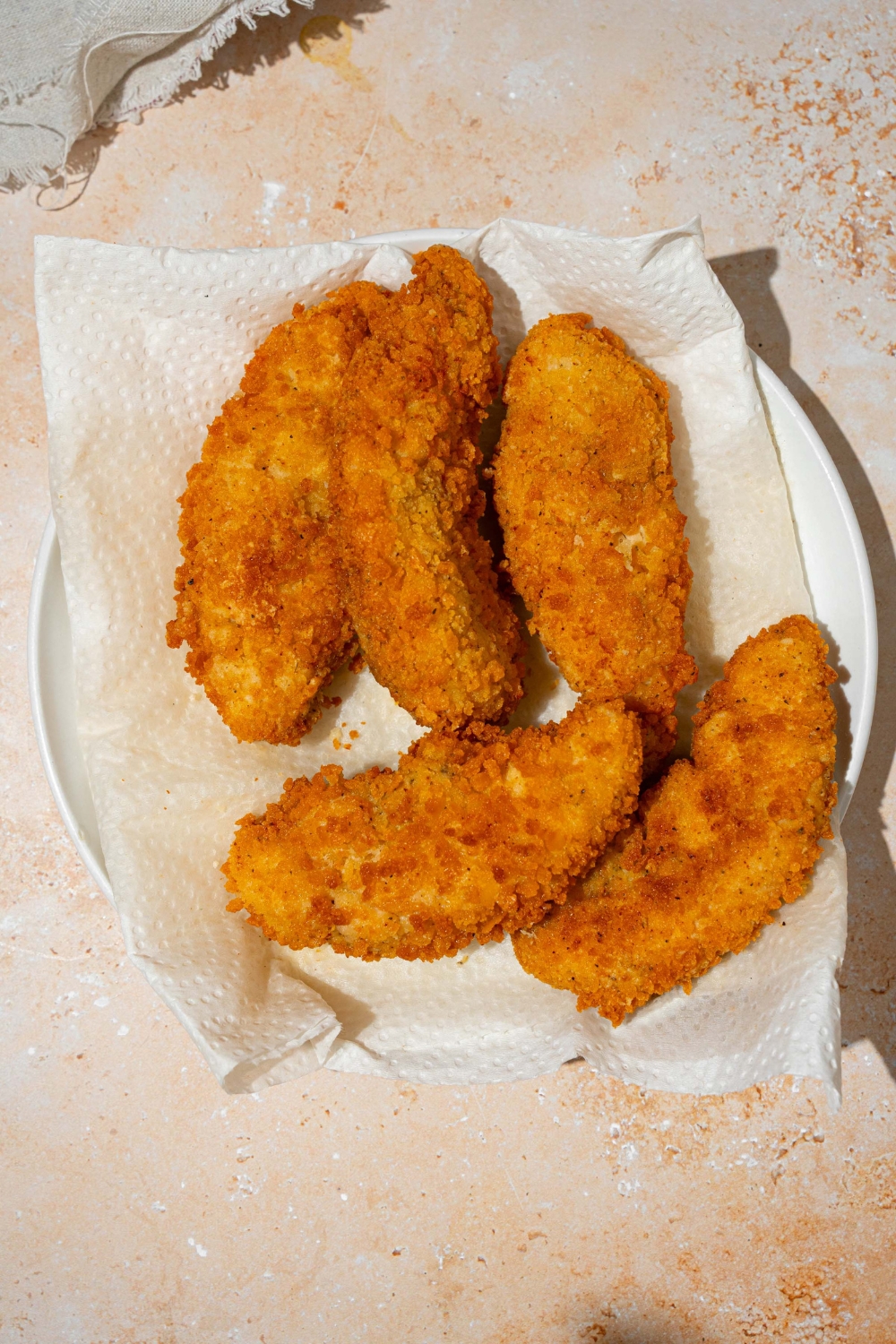 A white plate lined with parchment paper with fried chicken strips. The plate is on a tan counter.