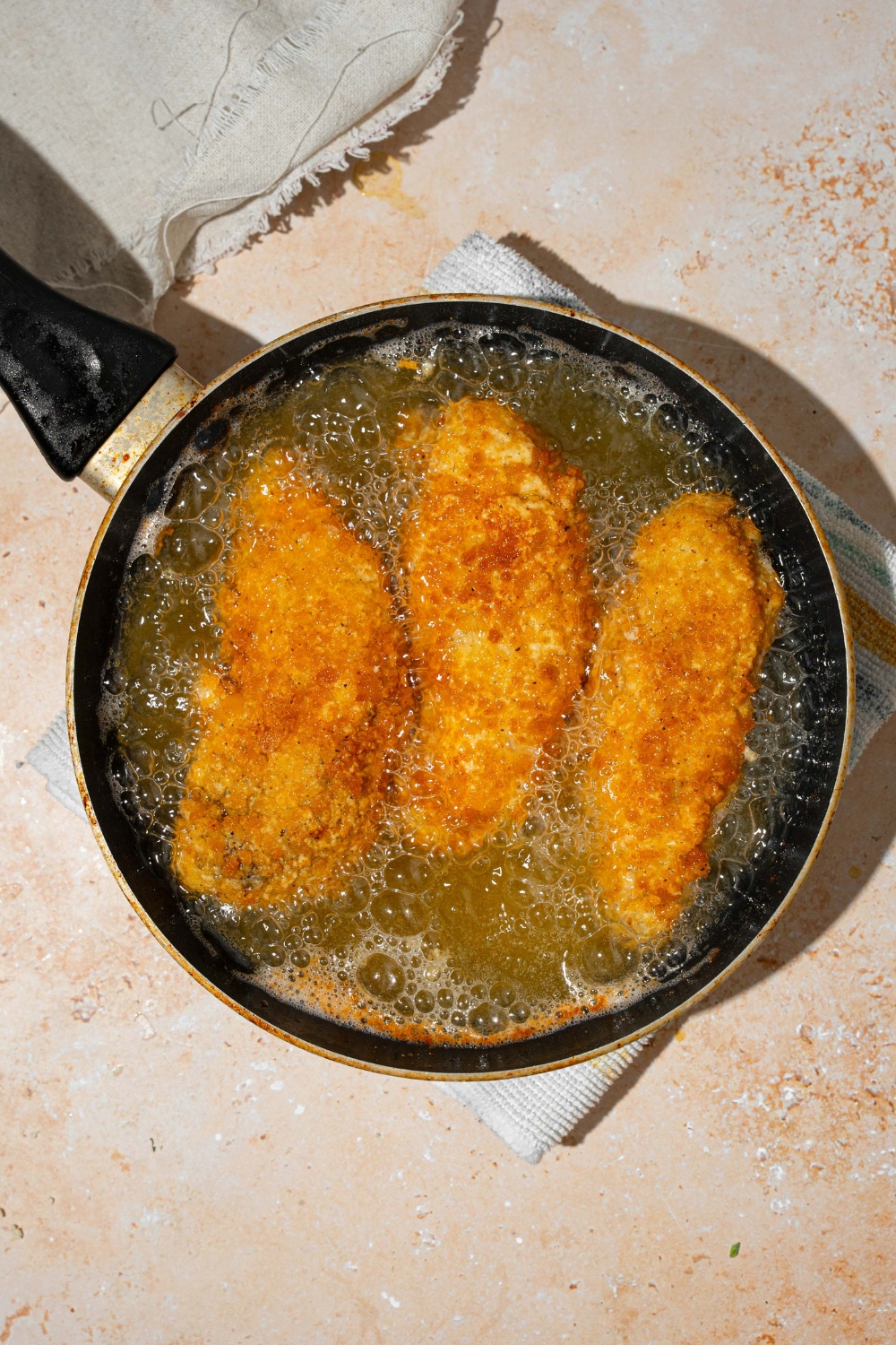 A skillet with three breaded chicken strips frying in oil. The skillet is on a tan counter with a white cloth napkin.