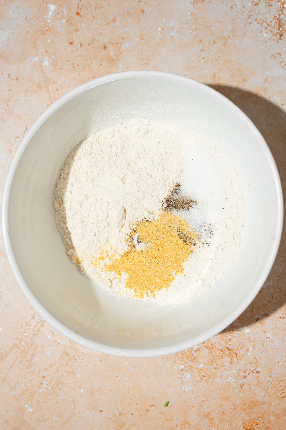 A white bowl with ingredients to make a flour mixture for breading including flour, garlic powder, salt, and pepper. The bowl is on a tan counter.