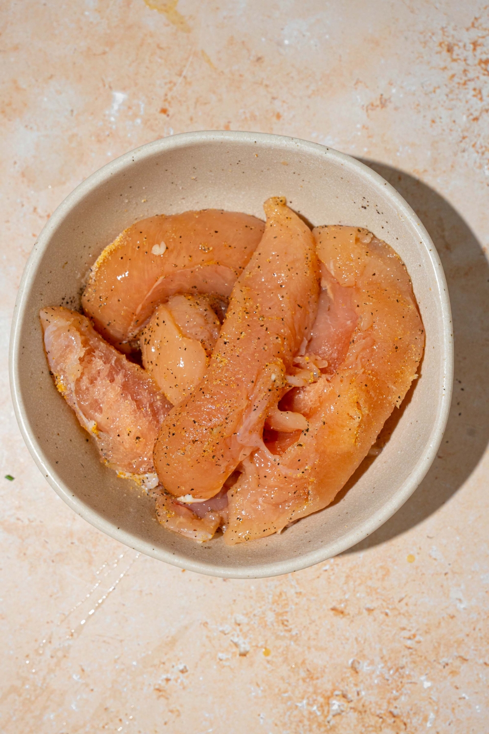 A white bowl with uncooked chicken strips seasoned with salt and pepper. The plate is on a tan counter.