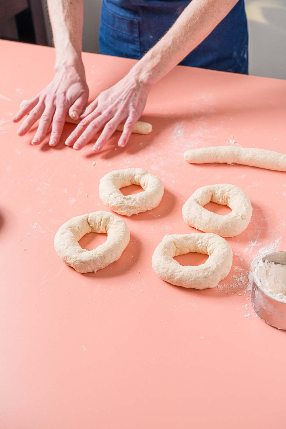 A pink counter with hands rolling out greek yogurt bagel dough next to several shaped unbaked greek yogurt bagels.