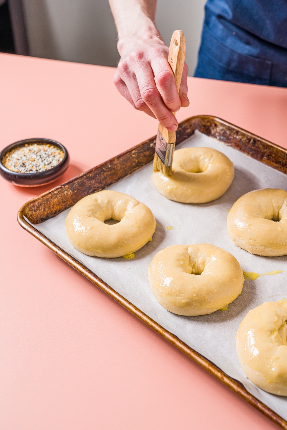 A baking sheet lined with parchment paper with several baked greek yogurt bagels being brushed with egg wash. The sheet is on a pink counter with a small bowl of everything bagel seasoning.