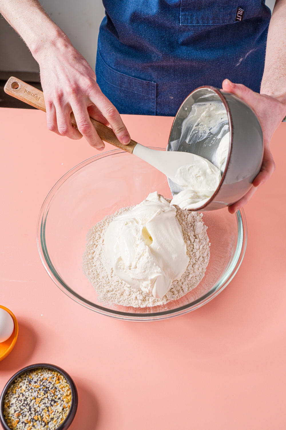 A glass bowl with ingredients being added to make greek yogurt bagels including greek yogurt and self rising flour. The bowl is on a pink counter.