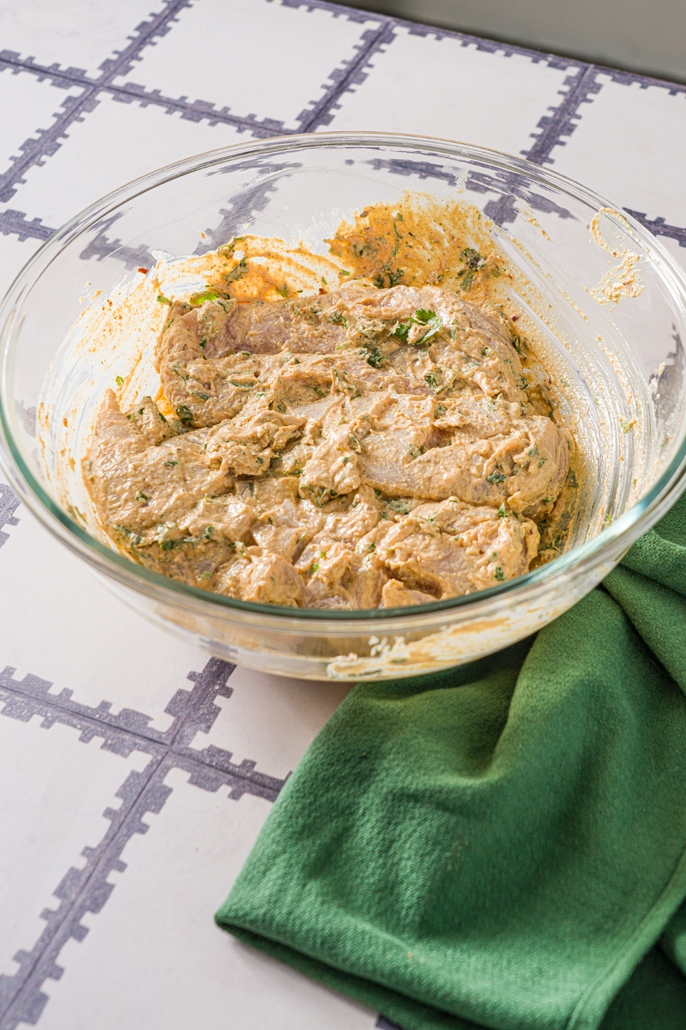A glass bowl with scored chicken breasts marinating in greek yogurt marinade. The bowl is on a tiled counter with a green cloth napkin.