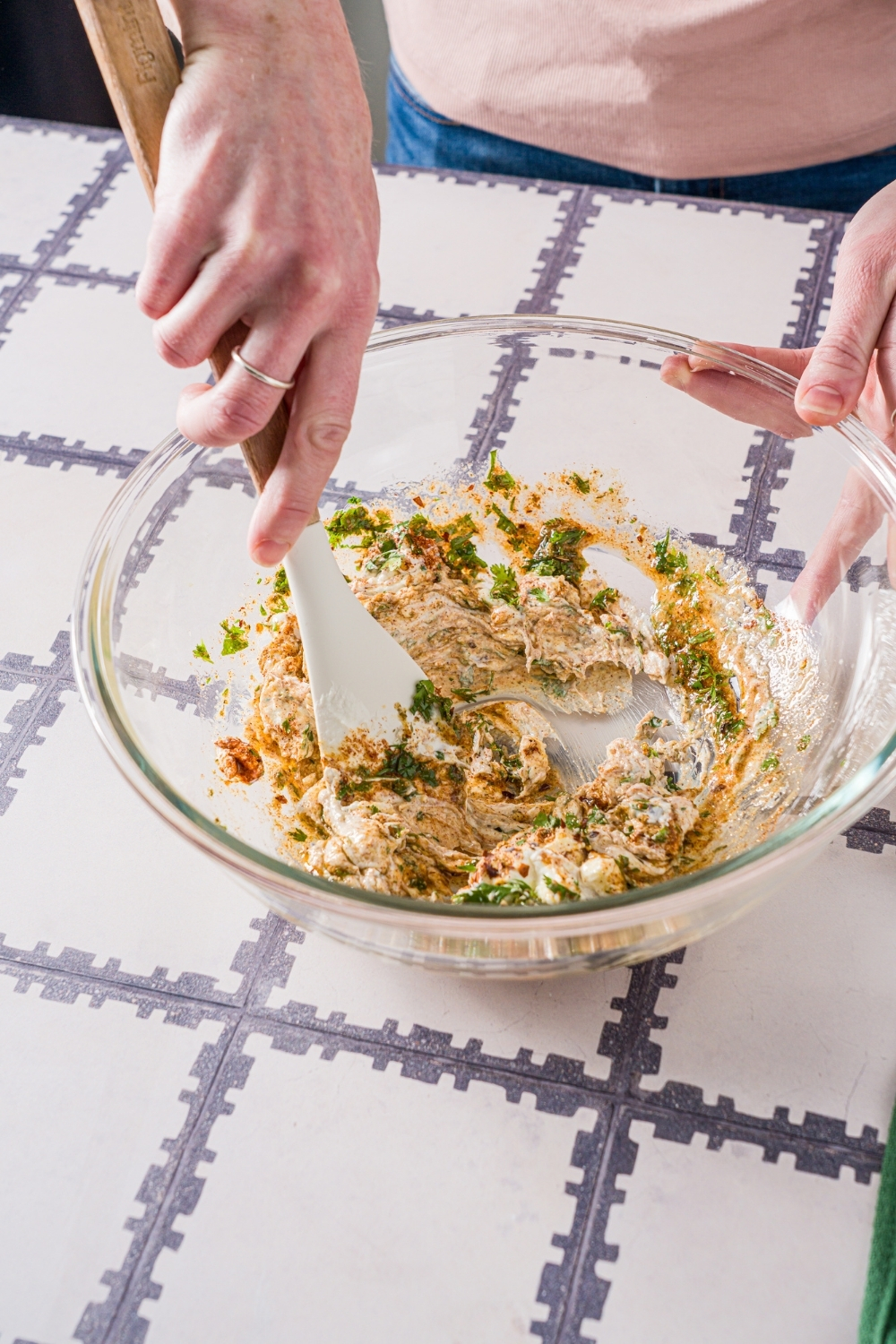 A glass bowl with a spatula mixing ingredients to make greek yogurt marinade including greek yogurt, olive oil, lemon juice, and seasonings. The bowl is on a tiled counter.