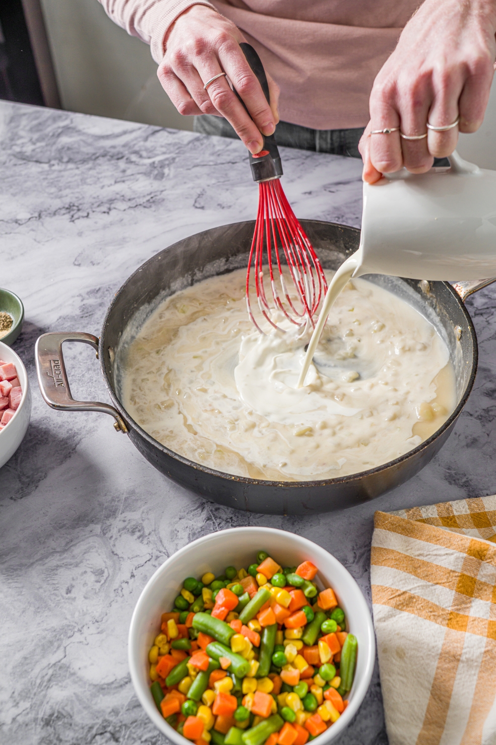 A skillet with heavy cream being whisked into a roux for pot pie. The skillet is on a marble counter with a small bowl of mixed vegetables.