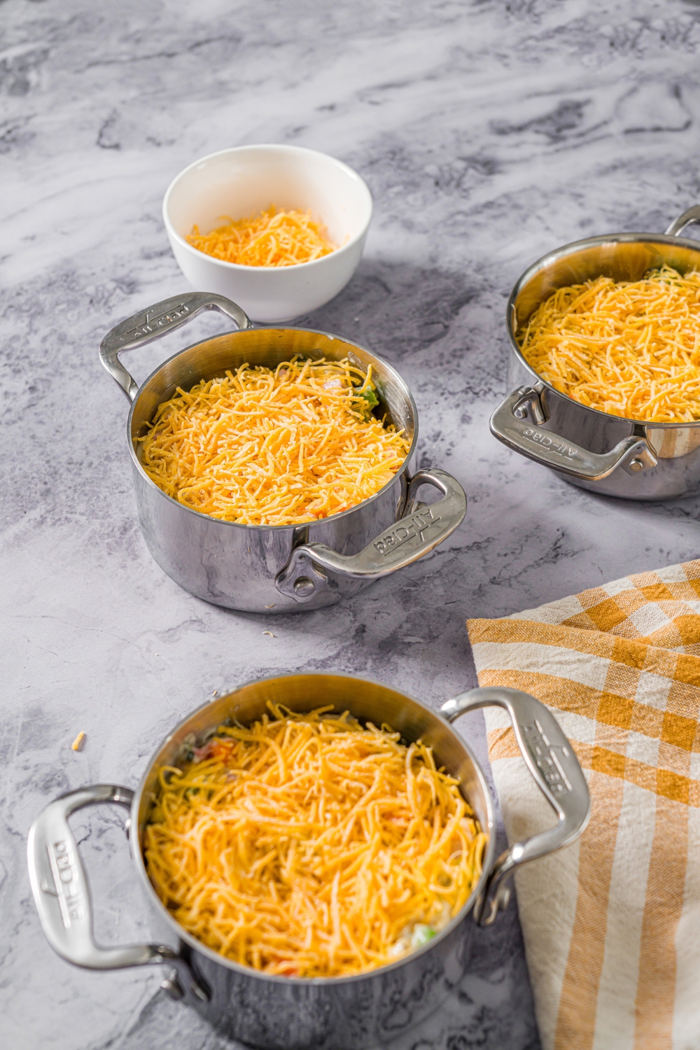 Three unbaked pie pots with pot pie filling topped with shredded cheese. The pots are on a marble counter with a yellow checkered napkin and bowl of shredded cheese.