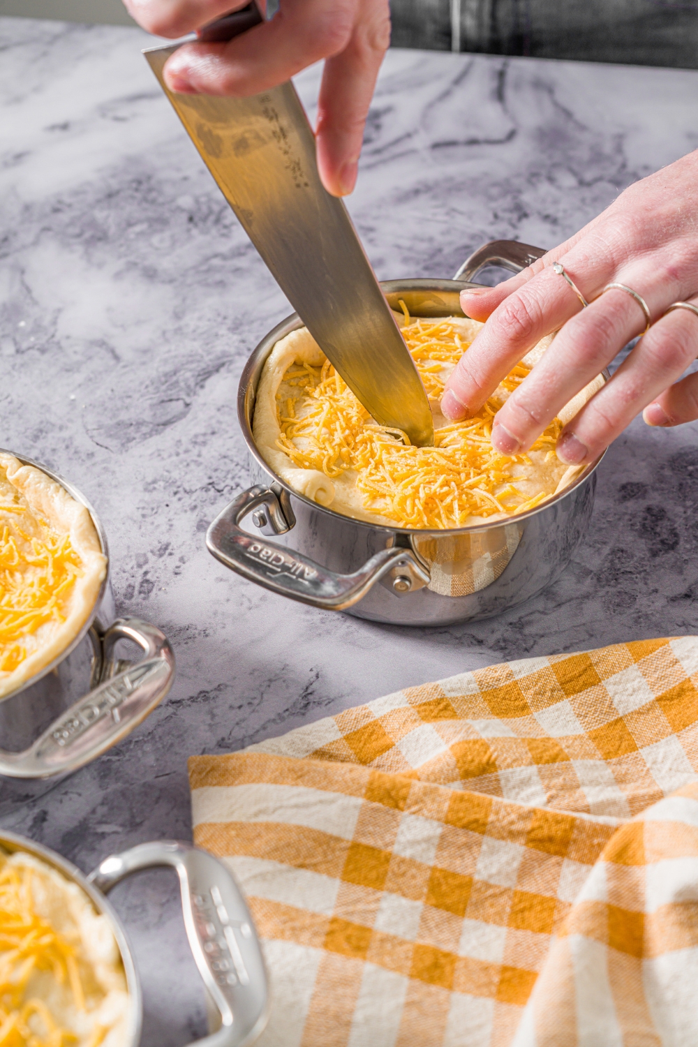 A pie pot with unbaked ham pot pie topped with dough and shredded cheese. A knife is slicing a slit into the pie. The pot is on a marble counter with a yellow checkered napkin.