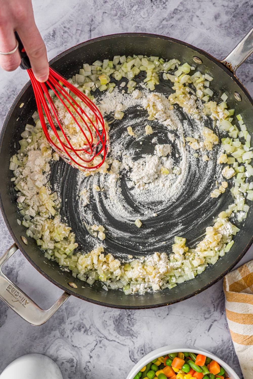 A skillet with sautéed onion with a whisk stirring flour to make a roux. The skillet is on a marble counter.