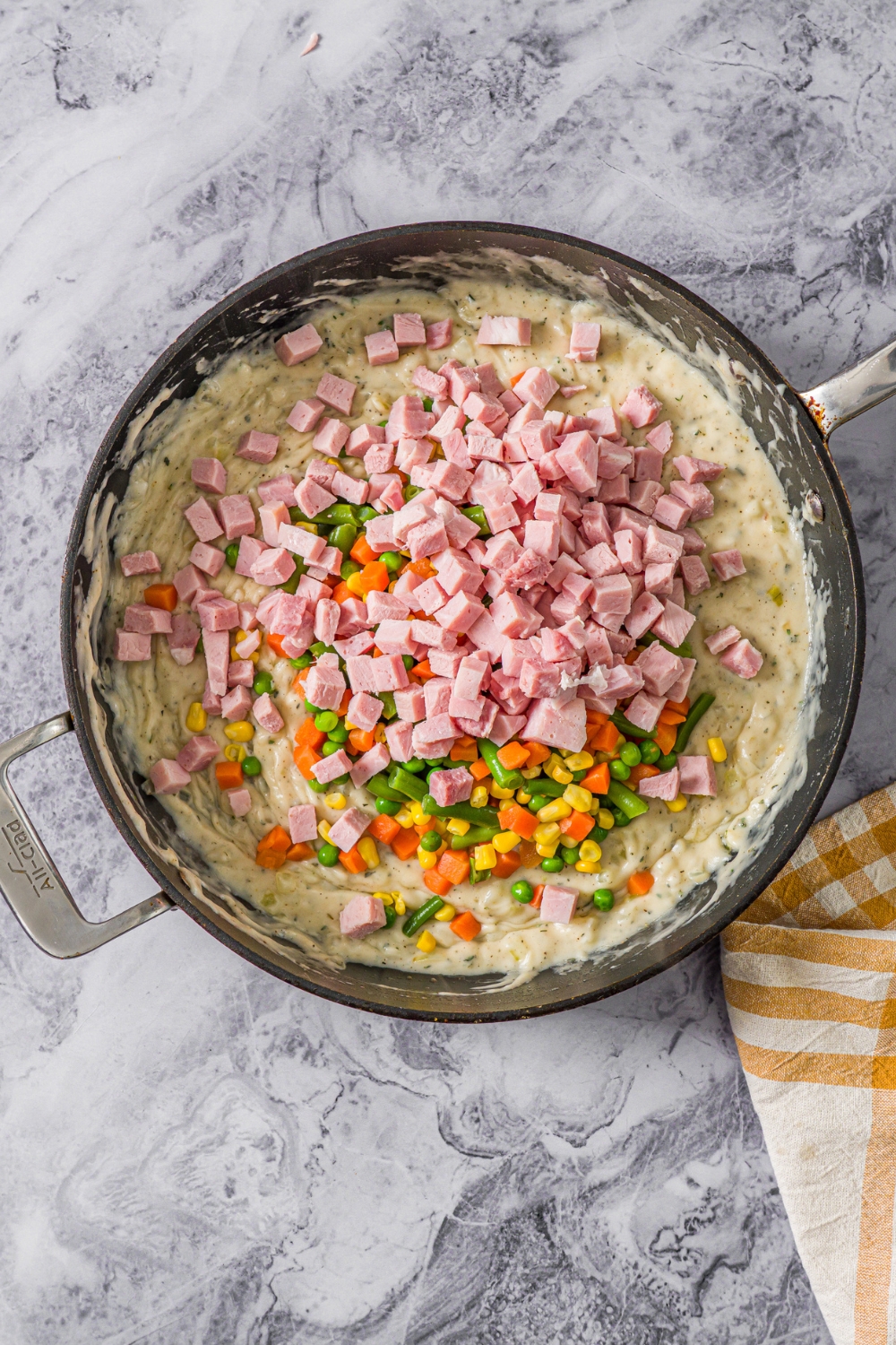 A skillet with ham and vegetables being added to a filling for pot pie. The skillet is on a marble counter.
