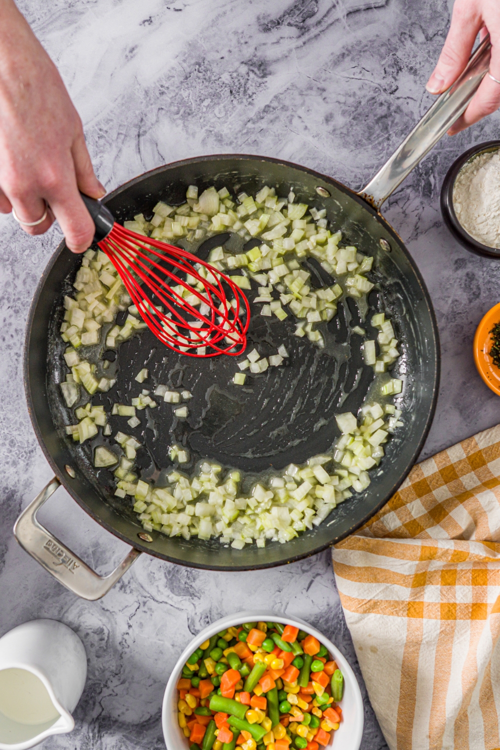 A skillet with a whisk mixing sautéed onion. The skillet is on a marble counter with a yellow checkered napkin.