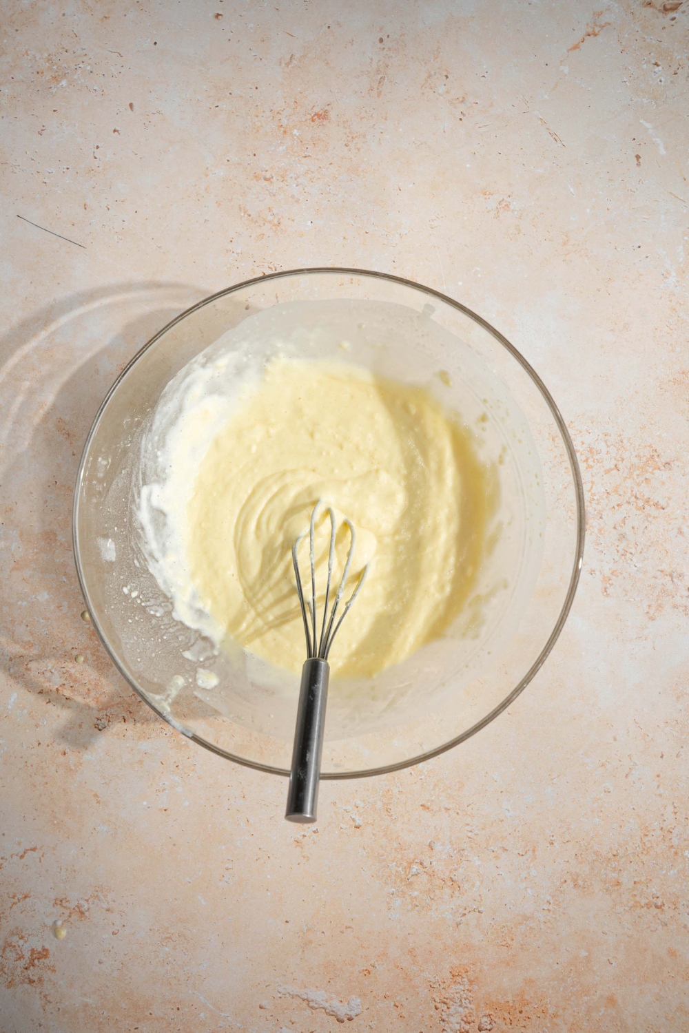A large glass bowl with a whisk stirring pancake batter with self rising flour. The bowl is on a tan counter.