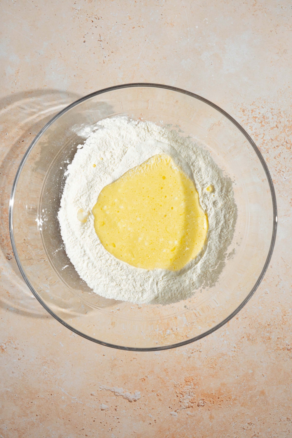 A large glass bowl with wet ingredients being added to dry ingredients for pancake batter. The bowl is on a tan counter.