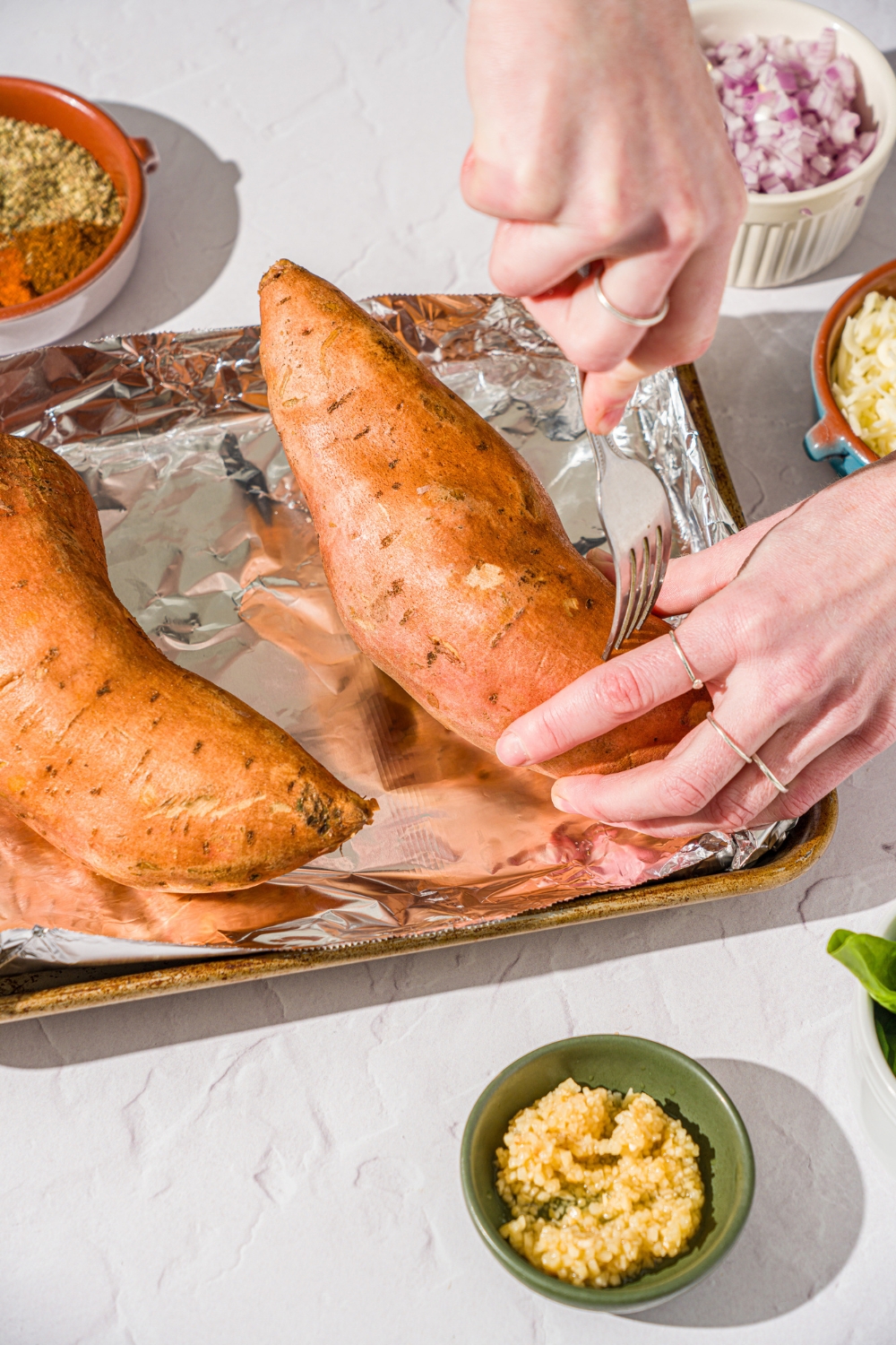 A baking sheet lined with foil with two sweet potatoes. A hand is piercing holes into the potato with a fork. The sheet is on a white counter with small bowls of ingredients.