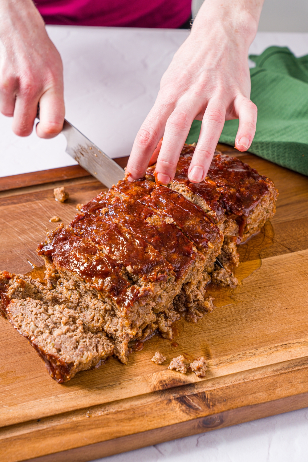 A wooden cutting board with a meatloaf made without eggs topped with a glaze. The meatloaf is being cut into slices. The board is on a marble counter with a green cloth napkin.