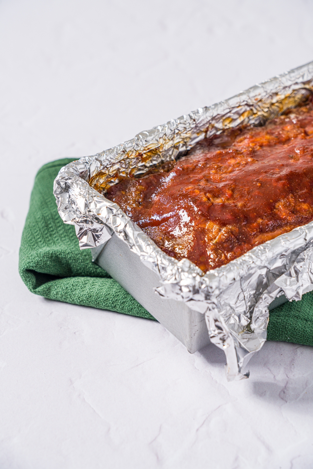 A bread pan lined with foil with baked meatloaf topped with glaze. The pan is on a marble counter with a green cloth napkin.