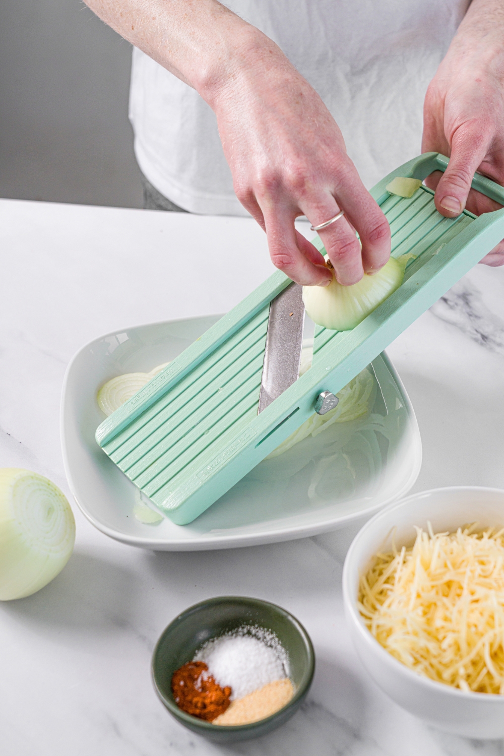 A mandolin slicing white onions into a white plate. The plate is on a marble counter with a bowl of shredded parmesan cheese and small bowl of seasonings.