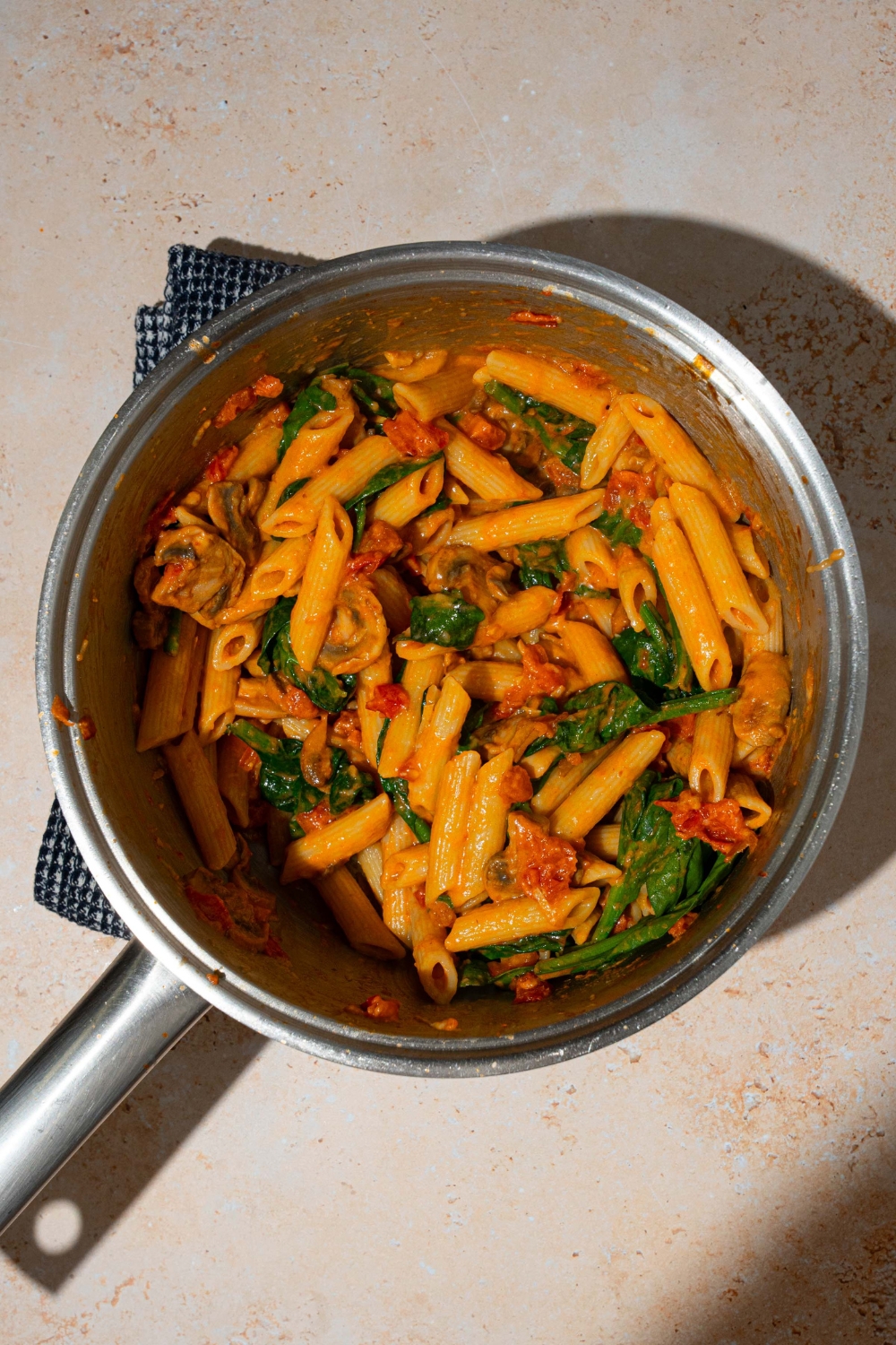 A pot with cooked penne rosa tossed with spinach and mushrooms. The pot is on a blue cloth napkin on a tan counter.