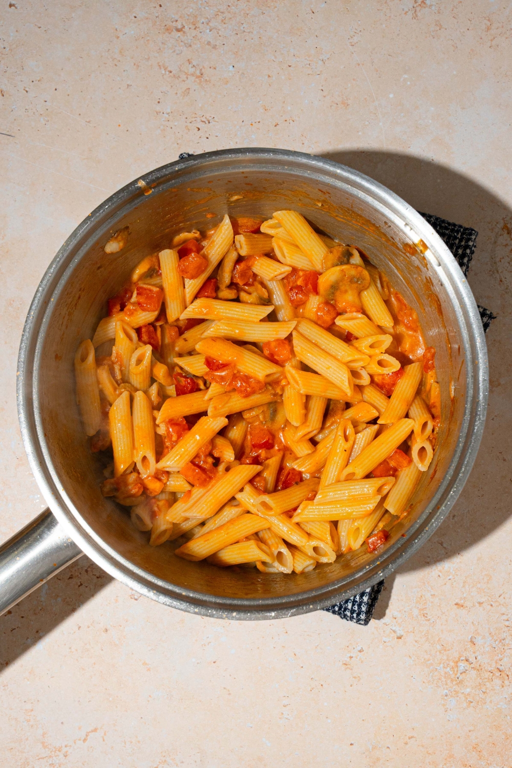 A pot with penne pasta tossed in penne rosa sauce. The pot is on a blue cloth napkin and tan counter.