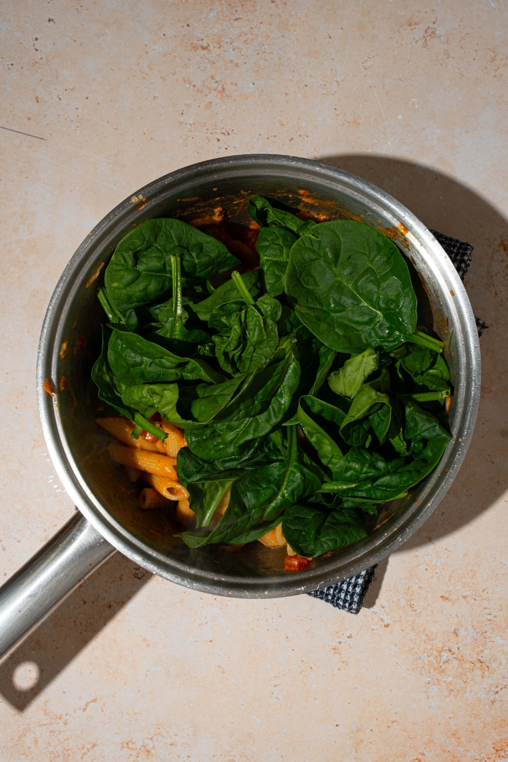 A pot with penne tossed in penne rosa sauce topped with spinach. The pot is on a blue cloth napkin on a tan counter.