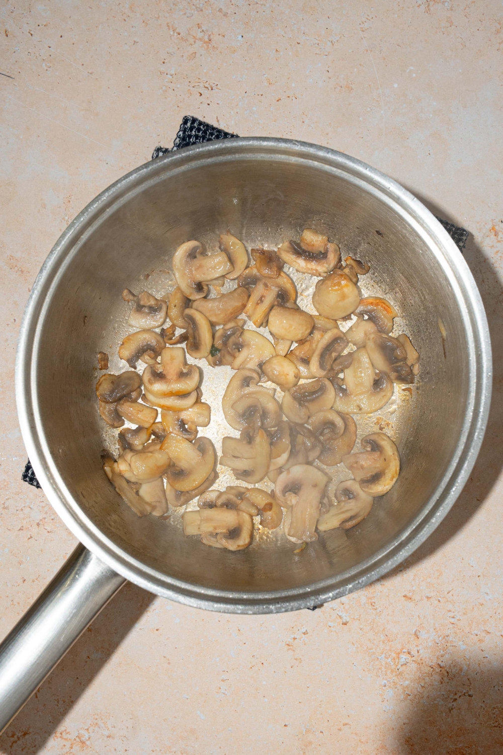 A pot with sliced mushrooms sautéing in oil. The pot is on a blue cloth napkin on a tan counter.