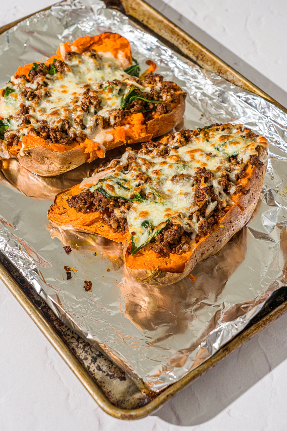 A baking sheet lined with foil with two baked loaded sweet potatoes topped with a beef mixture and melted cheese. The sheet is on a white counter.