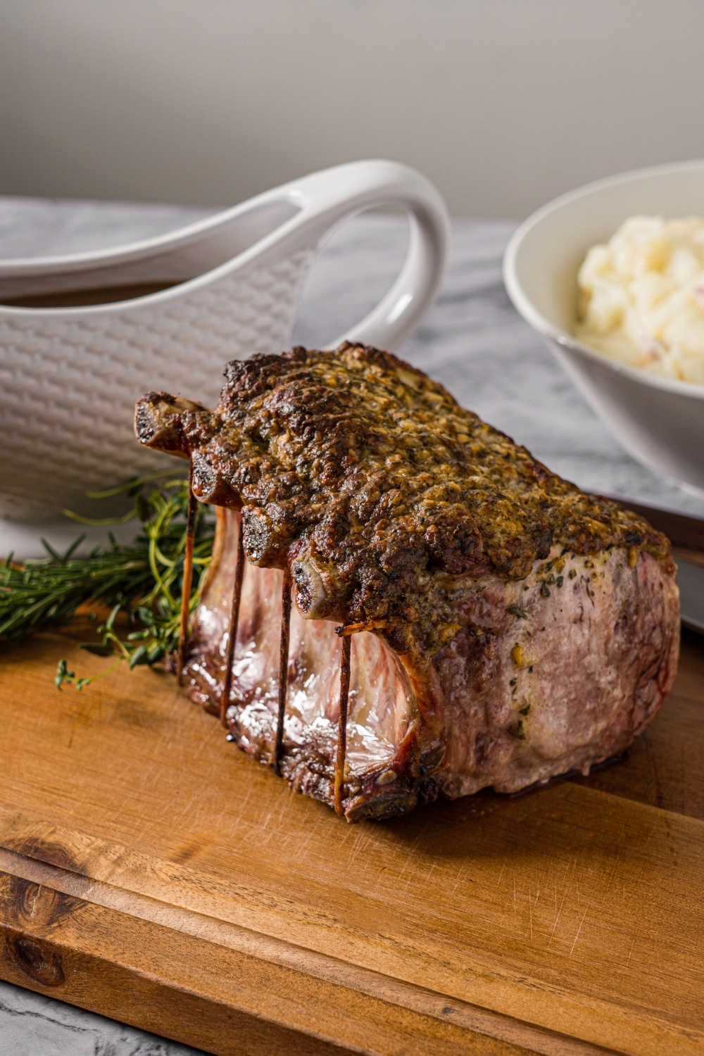 A whole rack of pork with a seasoned mustard mixture. The pork is on a wooden cutting board with a sharp knife and sprigs of rosemary. The board is on a marble counter with a bowl of mashed potatoes and gravy boat.
