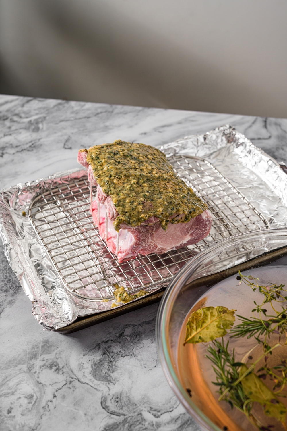 A baking sheet lined with foil and with a rack insert with an uncooked rack of pork topped with a seasoned mustard mixture. The sheet is on a marble counter.
