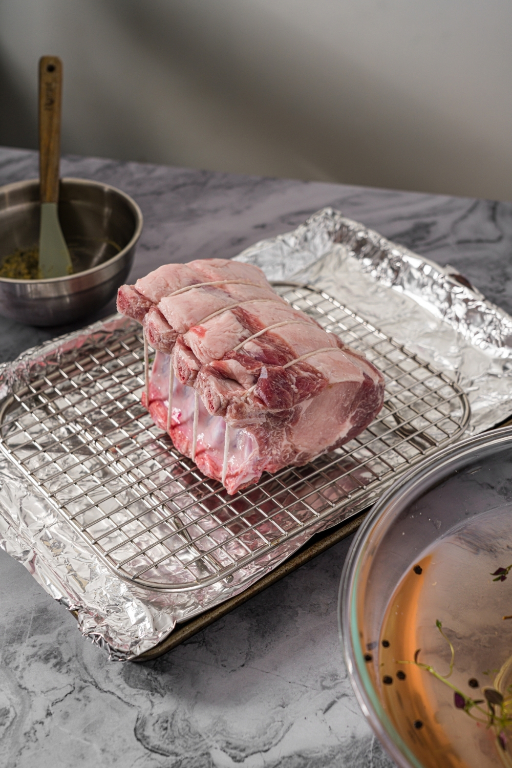 A baking sheet lined with foil and with a rack insert with an uncooked rack of pork. The sheet is on a marble counter with small bowl of toppings.
