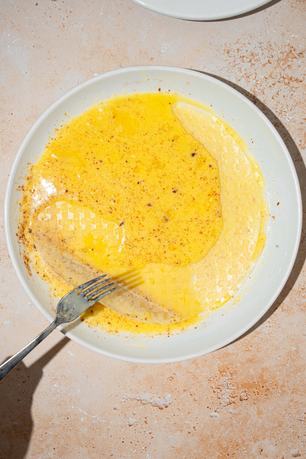 A white plate with an egg custard mixture with three sheets of rice paper. A fork is handling the sheets. The plate is on a tan counter.