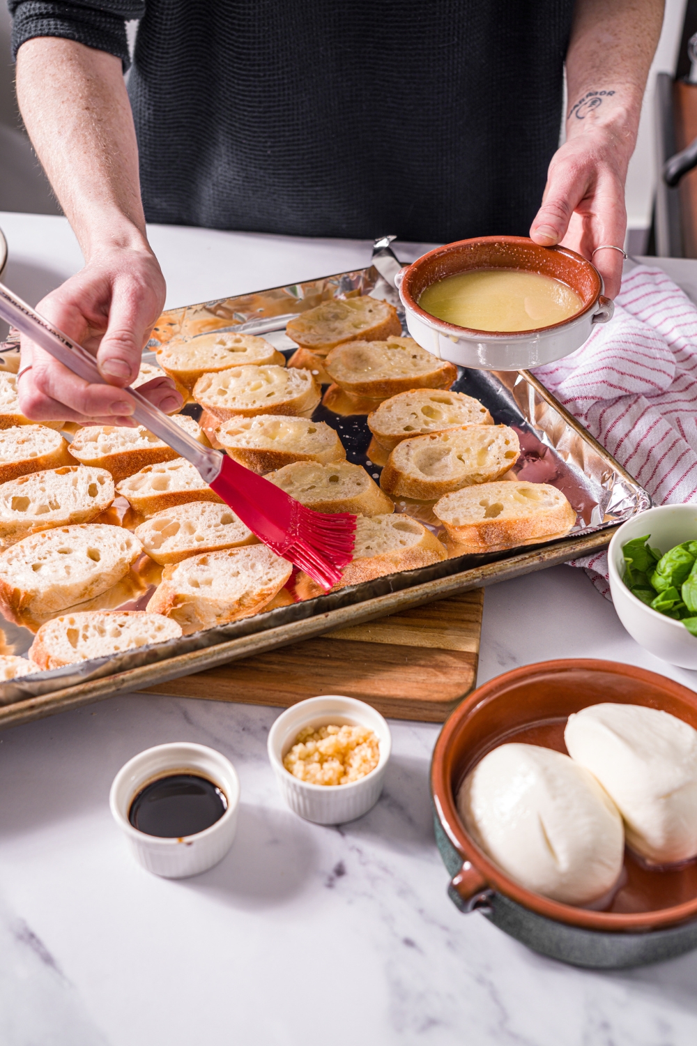 A baking sheet lined with foil with sliced baguette and a brush brushing garlic butter over the bread. The sheet is on a marble counter.