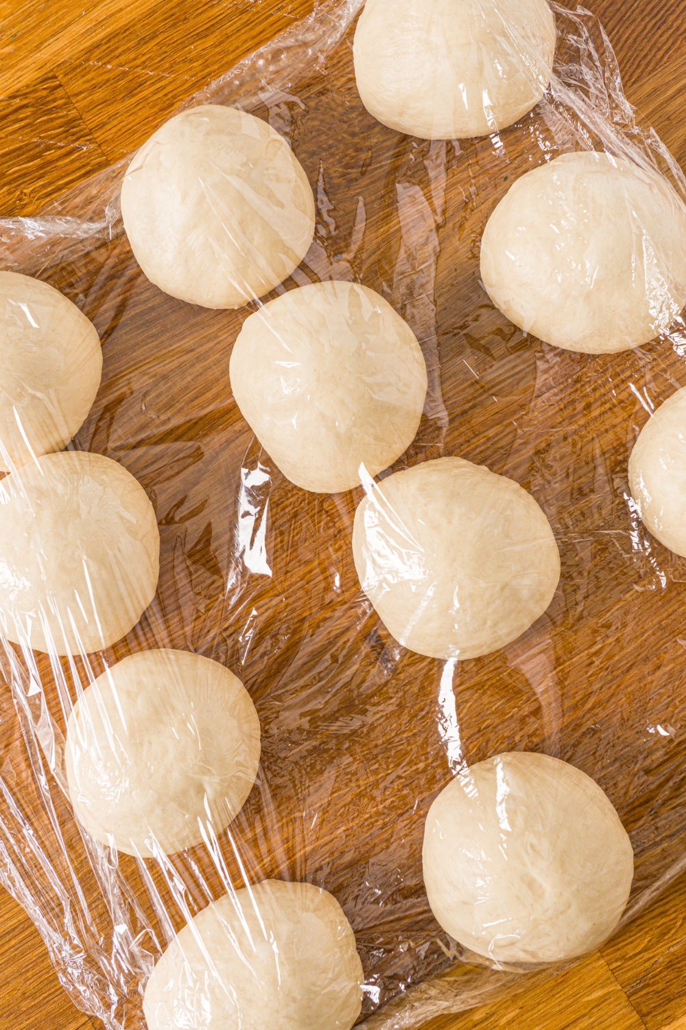 A wooden counter with several balls of salt bread dough with saran wrap over the balls.